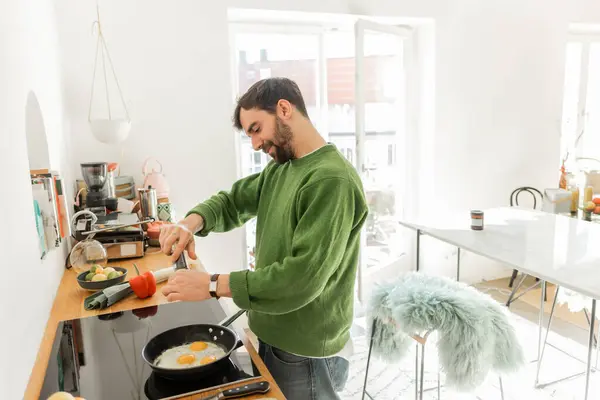 Smiling bearded man seasoning fried eggs with black peppercorns in mill while cooking breakfast — Stock Photo