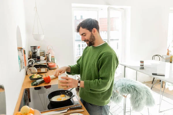Uomo barbuto in maglione verde condimento uova fritte durante la cottura della colazione al mattino — Foto stock