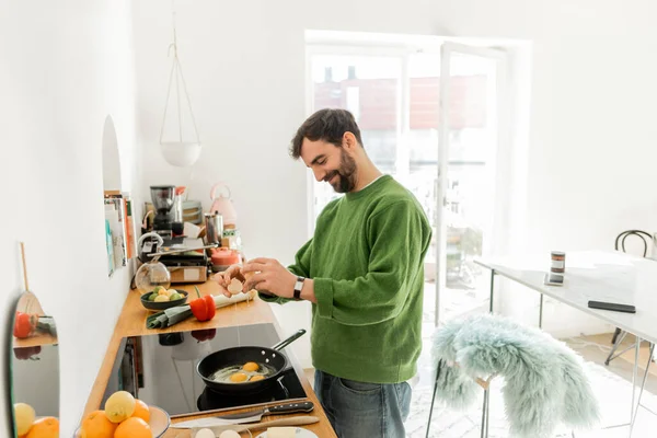 Allegro uomo barbuto in abiti casual versare l'uovo sulla padella mentre si cucina la colazione — Foto stock