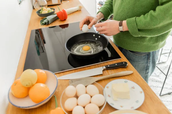 Vista cortada de homem tatuado segurando ovo perto da frigideira enquanto cozinha o café da manhã perto da manteiga — Fotografia de Stock
