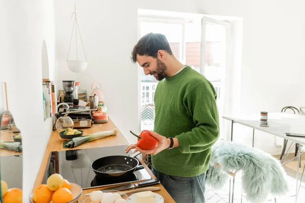 Hombre barbudo positivo sosteniendo pimiento cerca de la sartén mientras cocina el desayuno en casa - foto de stock