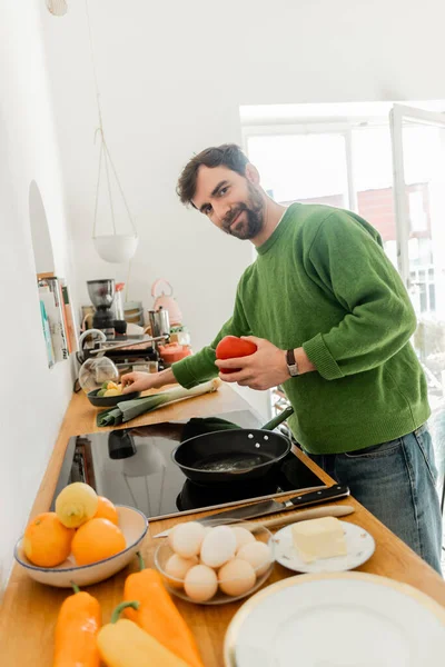 Smiling and bearded man in casual clothes looking at camera while holding bell pepper near eggs — Stock Photo