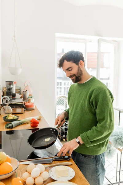 Радостный бородатый мужчина в зеленом свитере держит сковородку с маслом во время приготовления завтрака — стоковое фото