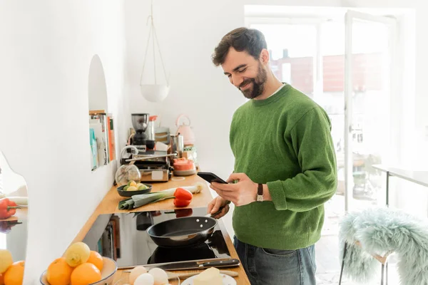 Positive and bearded man in casual clothes using smartphone while cooking breakfast — Stock Photo
