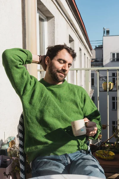 Hombre barbudo relajado con los ojos cerrados en jersey verde y jeans sosteniendo taza de café por la mañana - foto de stock
