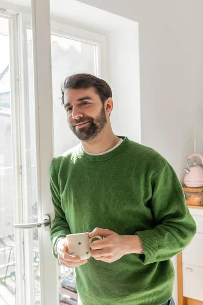 Positive bearded and brunette man in green jumper holding cup of coffee while standing near window — Stock Photo