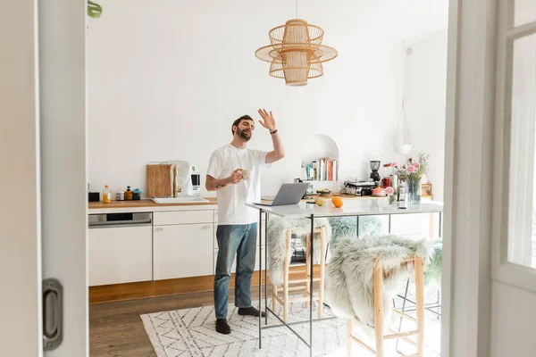 Bärtiger Mann in Freizeitkleidung mit Videotelefonie am Laptop und Kaffeetasse zu Hause — Stockfoto