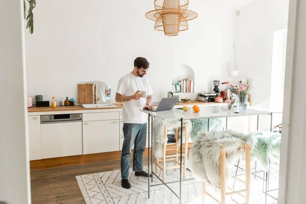 Freelancer barbudo en camiseta blanca y jeans sosteniendo taza de café y trabajando en el portátil en casa - foto de stock