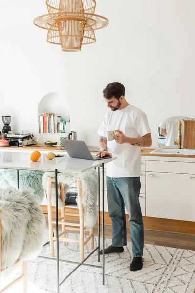 Freelancer en camiseta blanca y jeans sosteniendo taza de café y trabajando en laptop en casa - foto de stock