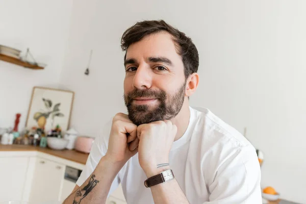 Retrato de homem barbudo e tatuado em camiseta branca olhando para a câmera, de mãos dadas perto do queixo — Fotografia de Stock