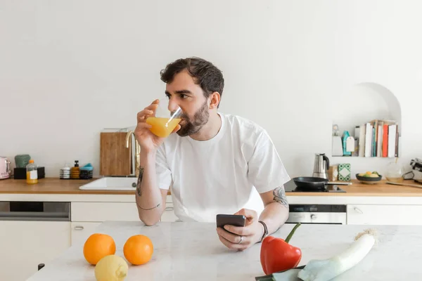 Hombre barbudo y tatuado en camiseta blanca bebiendo jugo de naranja fresco y usando smartphone - foto de stock