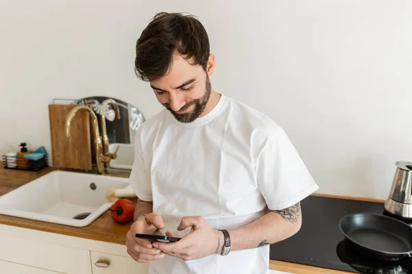 Uomo tatuato sorridente e barbuto in t-shirt bianca con smartphone mentre in piedi vicino al piano di lavoro — Foto stock