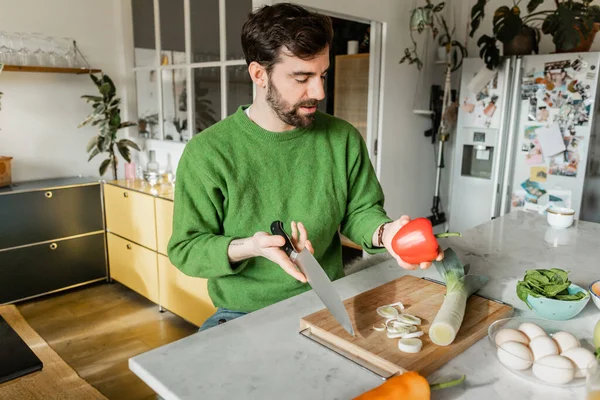 Бородатый татуированный мужчина в зеленом свитере держит нож и перец во время приготовления пищи на современной кухне — стоковое фото