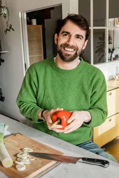 Hombre barbudo positivo en jersey verde sosteniendo pimiento fresco en la cocina en casa - foto de stock
