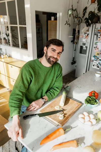 Positiver Mann im Pullover blickt in die Kamera, während er zu Hause in der modernen Küche neben frischen Lebensmitteln sitzt — Stockfoto