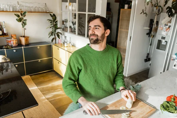 Entspannter und bärtiger Mann in grünem Pullover schaut weg, während er Messer und Lauch zu Hause hält — Stockfoto