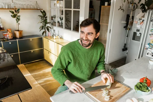 Веселый бородатый мужчина в зеленом свитере режет свежий лук-порей и смотрит в сторону, когда готовит дома — стоковое фото