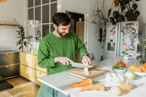 Positive bearded man in green jumper cutting fresh leek near orange juice in kitchen at home — Stock Photo