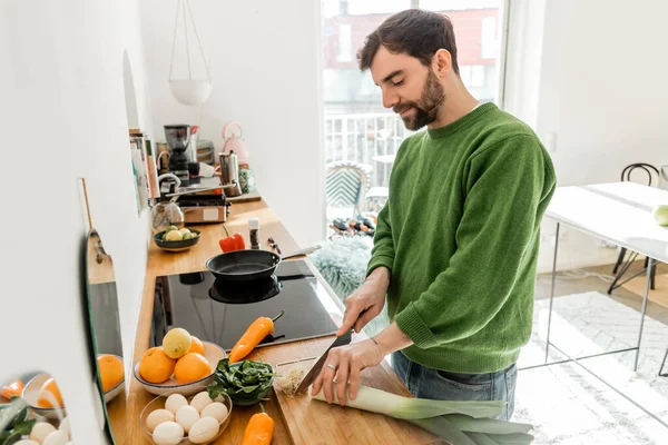 Giovane uomo barbuto in abiti casual taglio porro vicino uova e cibo fresco sul piano di lavoro a casa — Foto stock