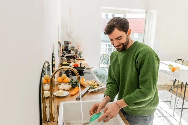 Веселый и татуированный бородатый мужчина в зеленом свитере и джинсах моет тарелку дома — стоковое фото