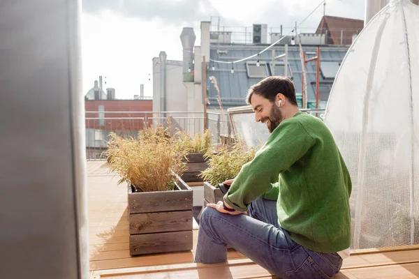 Uomo barbuto sorridente in auricolare e abiti casual utilizzando smartphone sul tetto terrazza della casa — Foto stock