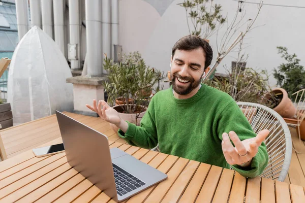 Hombre barbudo alegre en auriculares con videollamada en el ordenador portátil mientras trabaja en la terraza de la azotea - foto de stock