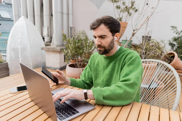 Bärtiger Unternehmer mit Kopfhörer und grünem Pullover hält Smartphone in der Hand und benutzt Laptop auf der Terrasse des Hauses — Stockfoto