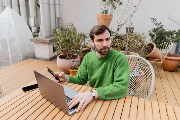 Freelancer barbudo en auriculares inalámbricos en puente verde usando smartphone y trabajando en laptop - foto de stock