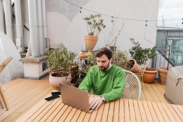 Empresario en auriculares inalámbricos utilizando el ordenador portátil mientras trabaja cerca de teléfonos inteligentes en la terraza de la casa - foto de stock