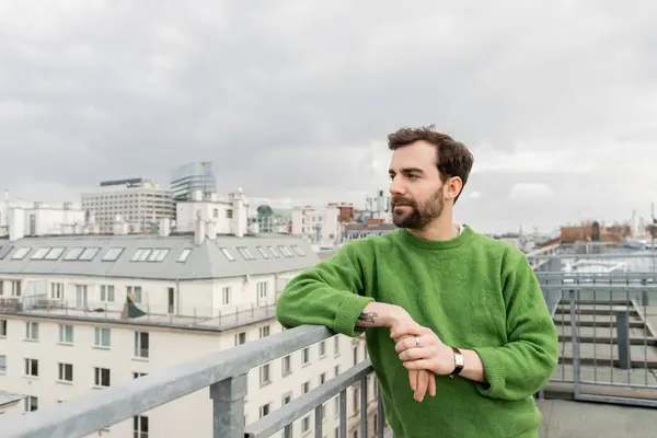 Tattooed man in green jumper looking away while standing on rooftop terrace in Vienna, Austria — Stock Photo