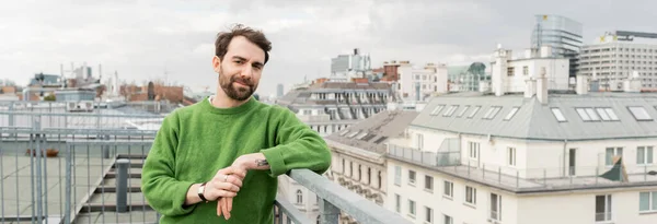 Tattooed man in green jumper looking away while standing on rooftop terrace in Vienna, banner — Stock Photo