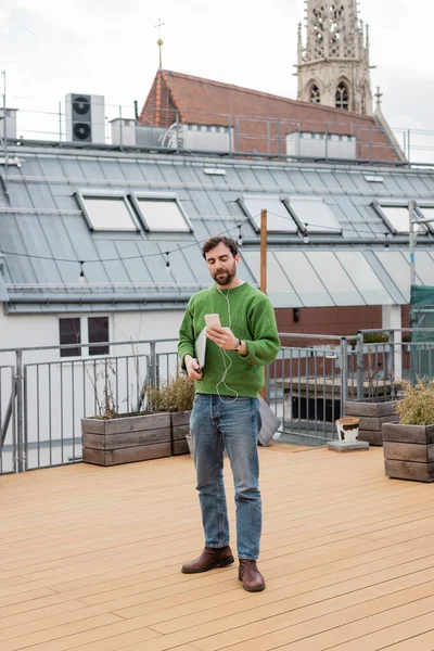 Freelancer con auriculares y smartphone y de pie en la terraza de la azotea de Viena - foto de stock