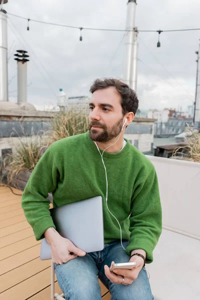 Freelancer barbudo em fones de ouvido segurando laptop no terraço em Viena, Áustria, trabalho remoto — Fotografia de Stock
