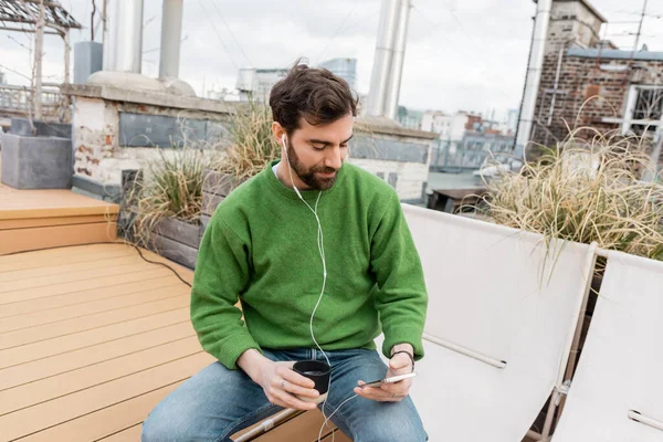 Bearded man listening music in earphones and holding cup of coffee on rooftop in Vienna, Austria — Stock Photo