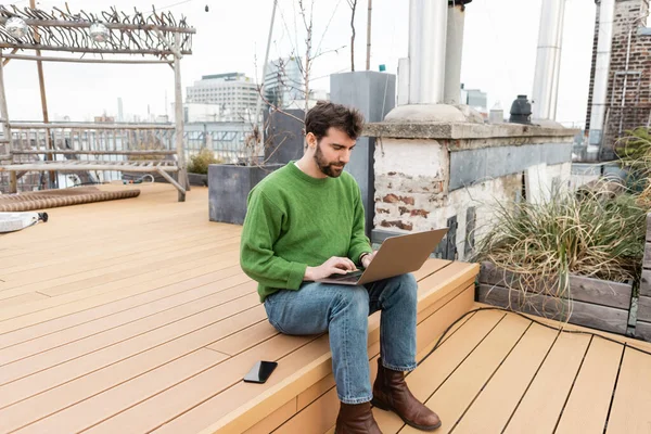 Bearded freelancer using laptop near smartphone while working on rooftop in Vienna, Austria — Stock Photo