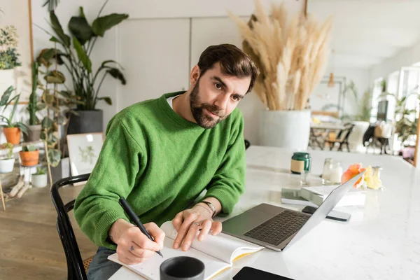 Brunette bearded freelancer in green jumper writing on notebook while working near devices on table — Stock Photo