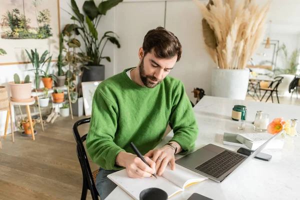 Bearded freelancer in green jumper writing on notebook near devices, cup of coffee and candles — Stock Photo
