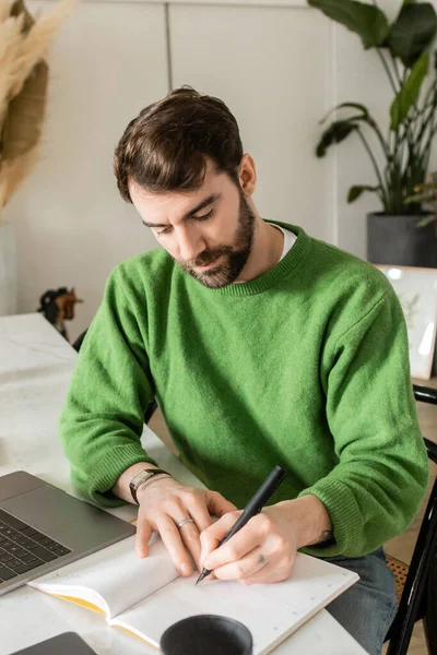 Bearded freelancer in green jumper writing on notebook near laptop, smartphone and cup of coffee — Stock Photo