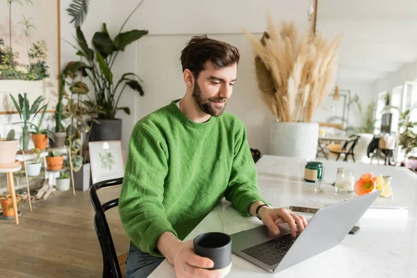 Freelancer feliz em jumper usando laptop e segurando xícara de café enquanto trabalhava em casa — Fotografia de Stock