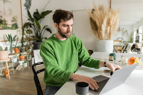 Freelance barbuto e bruna in maglione verde utilizzando il computer portatile e prendendo smartphone sul tavolo — Foto stock