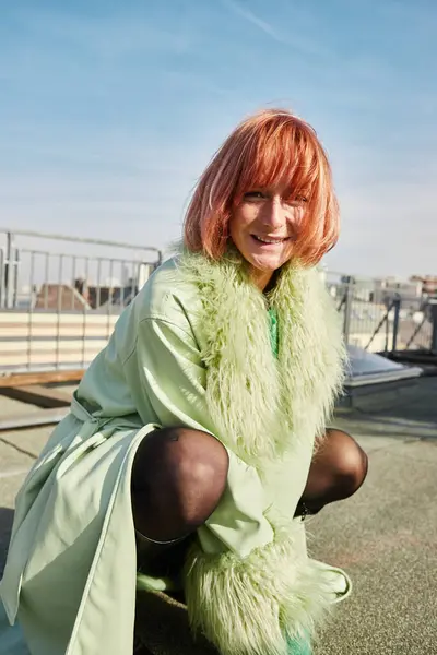 Mujer alegre y de moda sentada en las horcas y mirando a la cámara en la terraza de la azotea en Viena - foto de stock