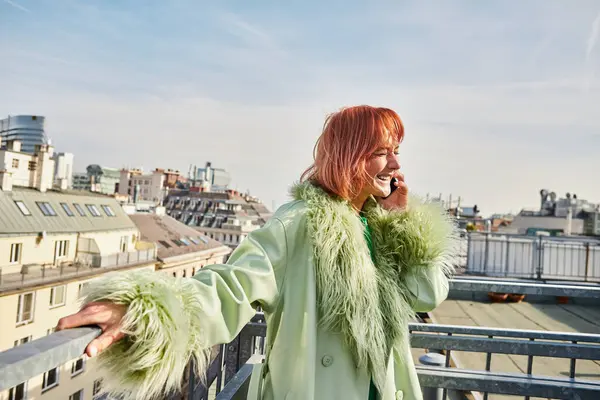 Радісна жінка в модному повсякденному вбранні розмовляє на мобільному телефоні на терасі на даху у Відні, Австрія — стокове фото