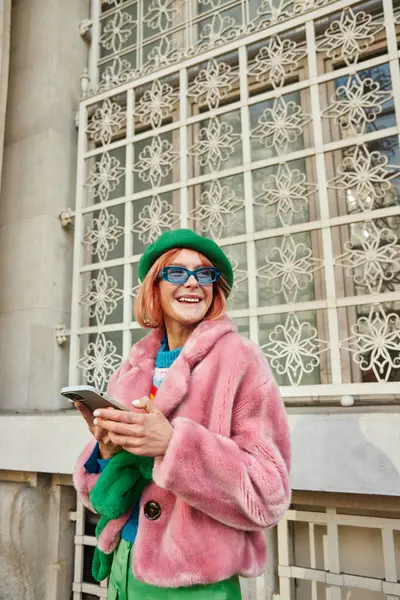 Carefree woman in sunglasses and stylish faux fur jacket using smartphone on urban street in Vienna — Stock Photo