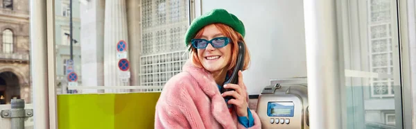 Mujer con estilo en gafas de sol y chaqueta de piel sintética sonriendo en cabina telefónica en la calle en Viena, pancarta - foto de stock