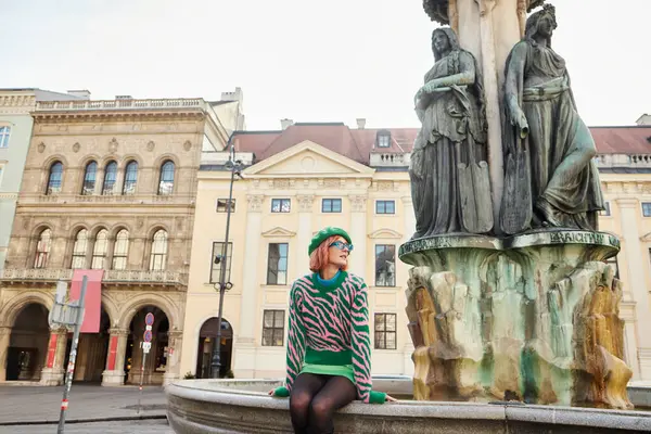 Donna in abbigliamento casual alla moda seduta vicino alla scultura e guardando lontano in strada a Vienna, Austria — Foto stock