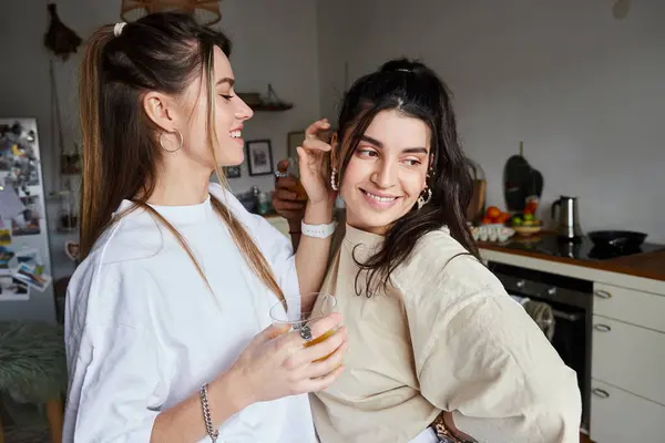 Happy young woman holding glass of fresh orange juice and flirting with girlfriend in kitchen, lgbt — Stock Photo