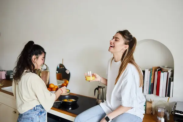 Happy young woman cooking breakfast near laughing girlfriend with orange juice, lgbt couple — Stock Photo