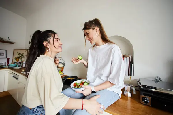 Happy young lesbian woman in 20s eating salad and looking at her girlfriend in kitchen, lgbt love — Stock Photo