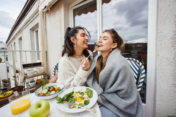 Happy young lesbian woman feeding her girlfriend while having breakfast on balcony, lgbt couple — Stock Photo