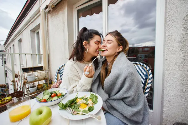 Sonriente joven lesbiana alimentar a su novia mientras desayuna en balcón, lgbt pareja - foto de stock